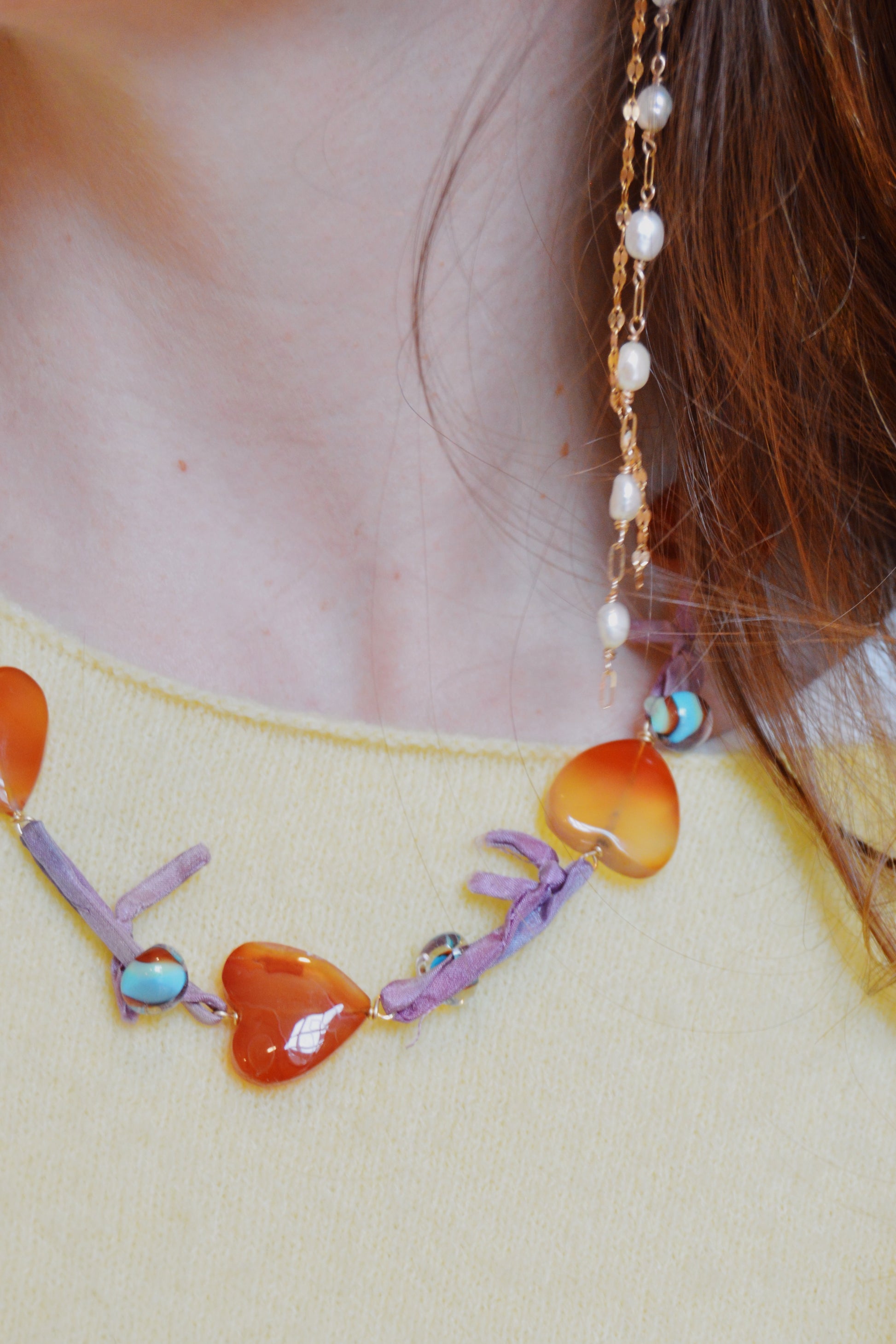 A close-up of a person in a yellow sweater models the Silk and Gemstone Heart Layer—a necklace with orange heart beads, purple accents, and gemstone hearts—plus a long gold earring with pearls. Brown hair partly covers their ear.