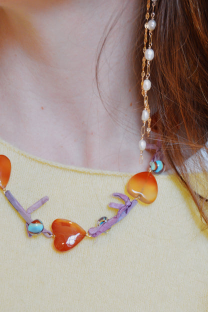 A close-up of a person in a yellow sweater models the Silk and Gemstone Heart Layer—a necklace with orange heart beads, purple accents, and gemstone hearts—plus a long gold earring with pearls. Brown hair partly covers their ear.