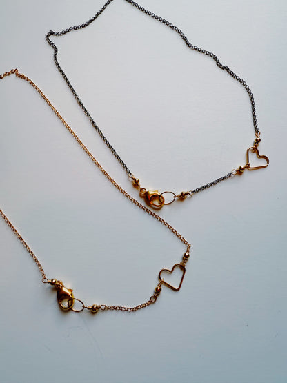 The Clasp in Front Layer features two delicate necklaces: a 16" oxidized sterling silver chain and a 14K gold filled heart pendant, both adorned with simple heart charms and small clasps on a white background.