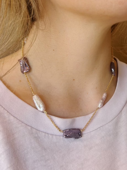 A close-up of a person wearing the Saturday Layer gold chain necklace with mixed rectangular purple and white pearls, paired with a pale lavender crew-neck top.