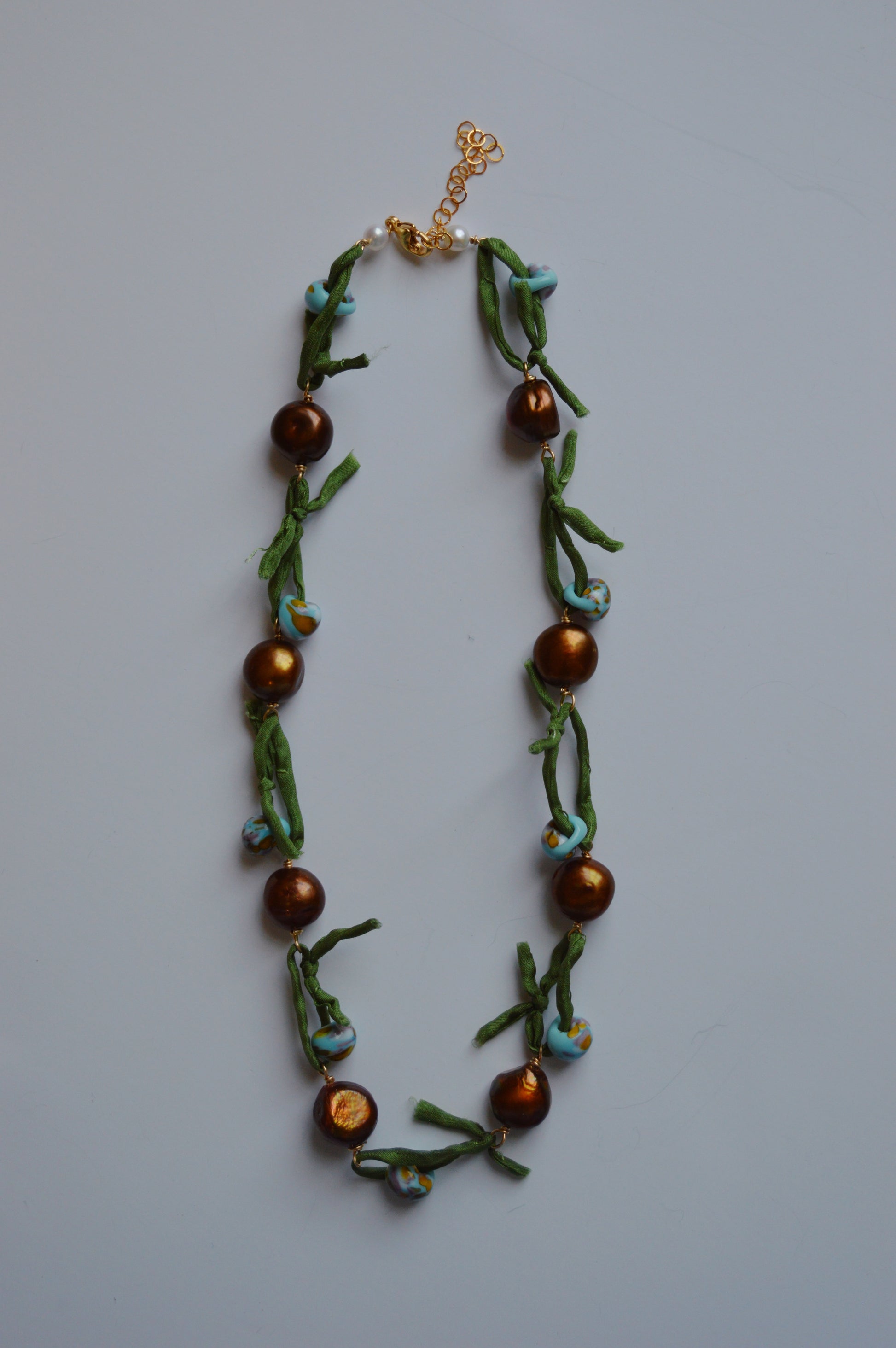 The Silk and Pearl Layer- Green Dunes is a handmade glass bead necklace with large round brown beads, small blue flower-shaped beads, and green ribbon accents linked by a gold chain and clasp, displayed on a white background.