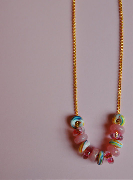 The Valentine Lots of Donuts Layer is a gold chain necklace featuring colorful, round beads in pink, blue, yellow, and white, showcased on a pale pink background.