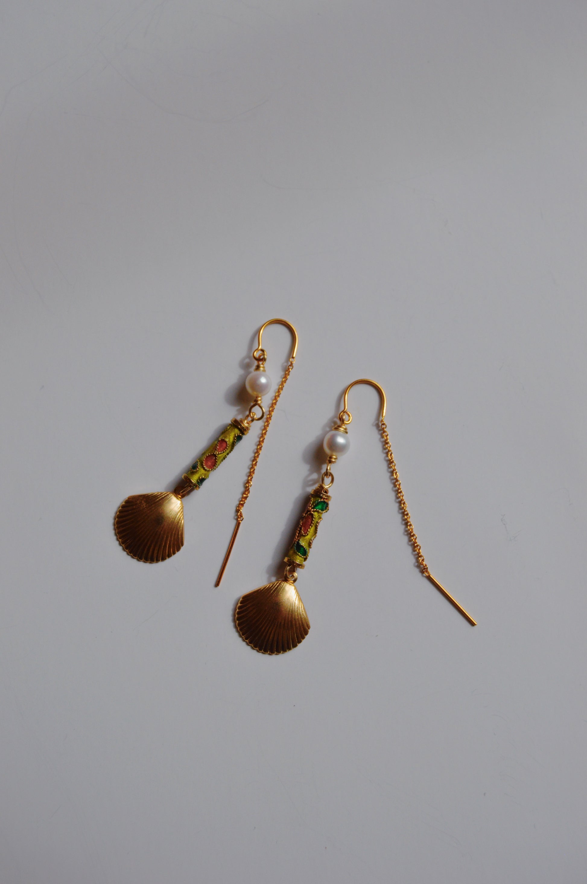 The Cloisonne Shell Threaders are gold drop earrings featuring scallop shell-shaped pendants, cultured freshwater pearls, and green and red detailing—handcrafted artisan jewelry that shines on a plain white background.