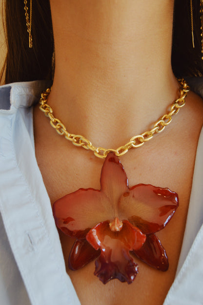 Wearing the Large Orchid Necklace, a 14k gold filled chain with a bold red preserved orchid pendant, paired with a light blue collared shirt. Only the neck and part of the shirt are shown.