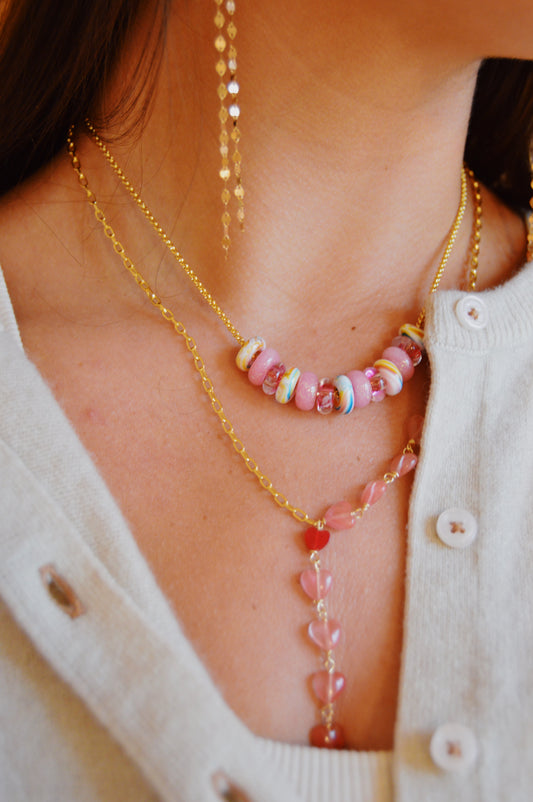Close-up of a person wearing the Valentine Gemstone Heart Lariat layered with gold necklaces and long gold earrings, paired with a light-colored buttoned top. One necklace features pink and multicolored beads, another has pink heart-shaped beads.