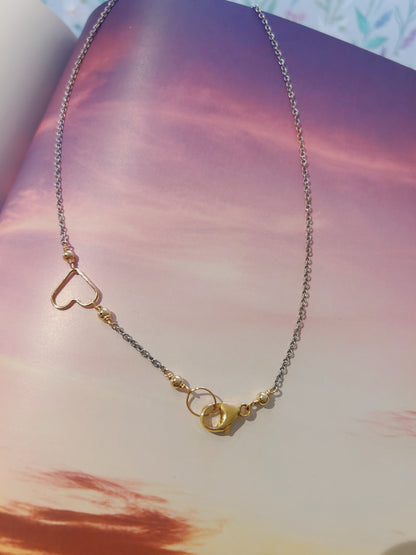 The Clasp in Front Layer necklace features a 14K gold filled heart outline with small round beads and mixed metals, hanging from a 16-inch oxidized sterling silver chain. Displayed on an open book with a pink and purple sunset sky.