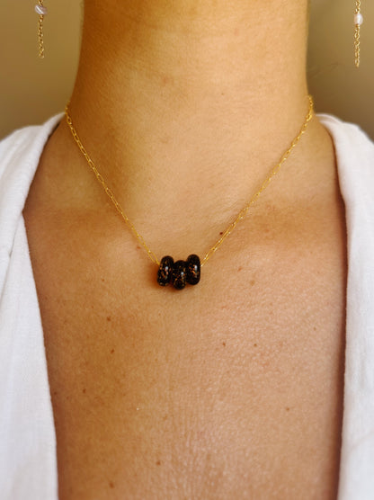 A close-up shows a person in a white top wearing the Dainty Donut Layer—a delicate 14K gold chain necklace with three dark, textured beads centered and a secure gold filled clasp—against a softly blurred background.