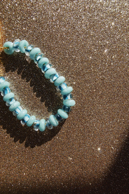 The Donut Bracelet, featuring blue beads and gold filled findings, is displayed on a sparkling glittery brown surface where sunlight casts a shadow and highlights the shine of both the bracelet and background.