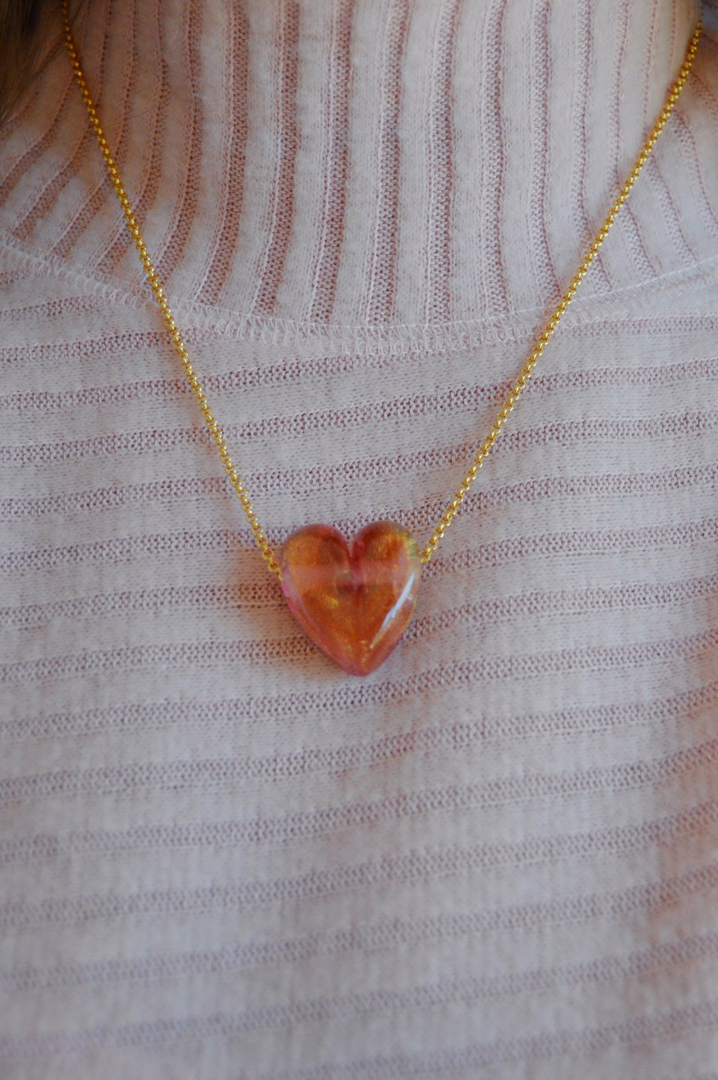 A person wears the Glass Heart Necklaces- Reds & Oranges, featuring a handmade lampwork glass heart pendant on a gold-filled chain, shown against a white ribbed turtleneck sweater.