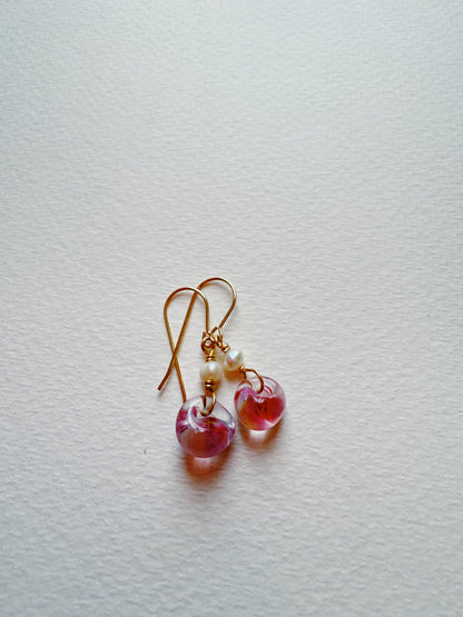 The Bubble Minis are gold hook earrings with delicate glass beads, small white accents, and translucent pink heart-shaped charms, shown on a textured white surface.