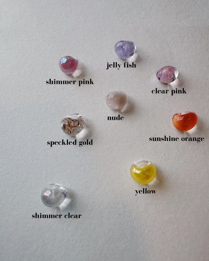 Seven small, round Bubble Drops—handblown glass beads in shimmer pink, jelly fish, clear pink, nude, speckled gold, sunshine orange, yellow, and shimmer clear—are arranged on a white surface.