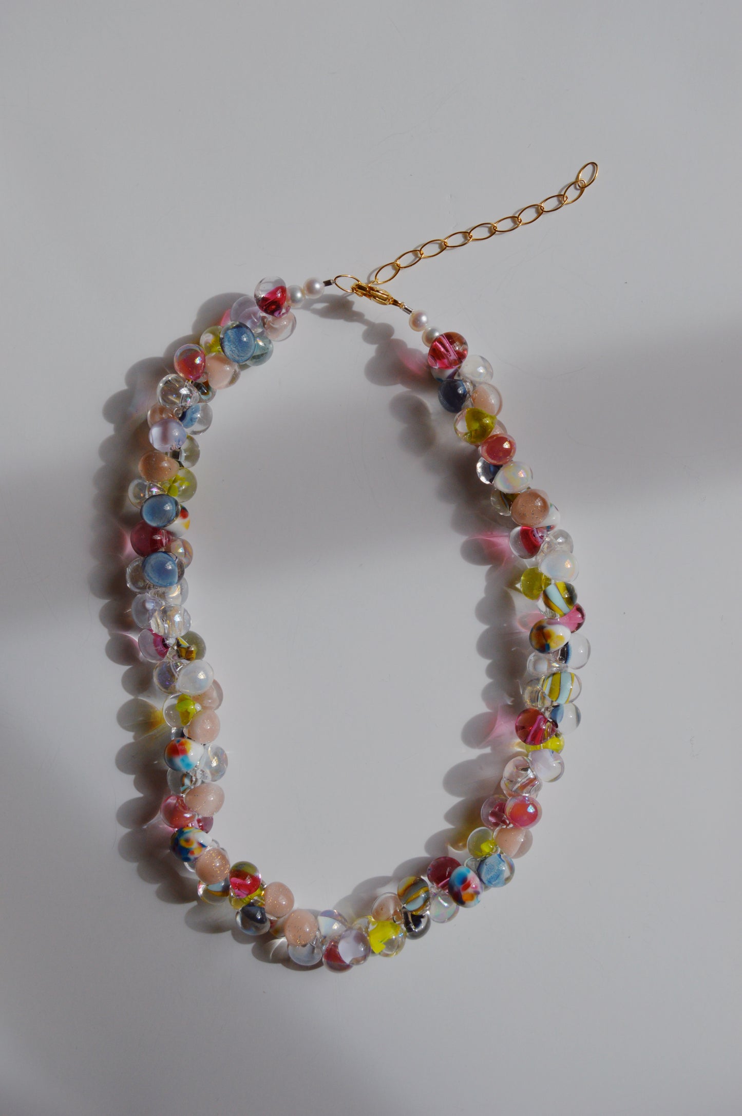 The Rainbow Bubble Glass Necklace features handmade lampwork glass beads in taffy colors, with pink, yellow, blue, and clear round beads arranged on a gold chain, displayed against a white background.