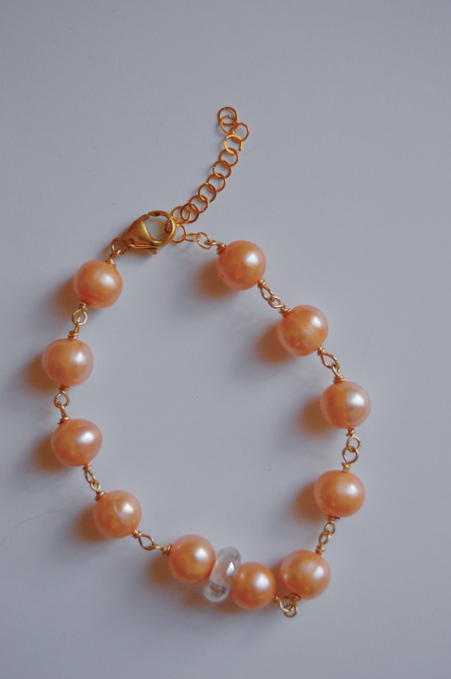 The Pearl and Glass Bracelet showcases a 14k gold-filled chain, champagne-colored freshwater pearls, and a handmade lampwork glass bead, displayed in a partial circle on a white background.