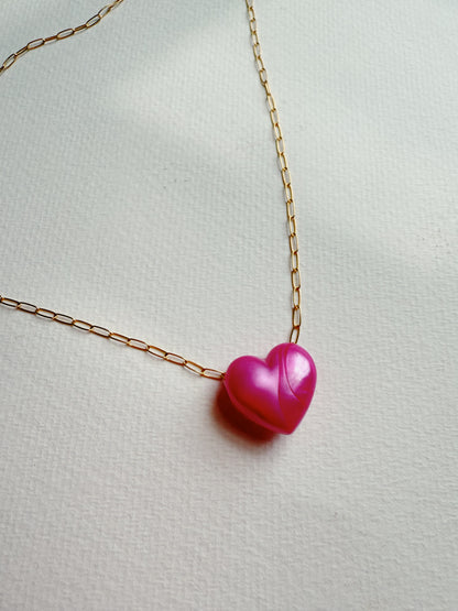 The Acrylic Heart Necklace features a shiny pink acrylic heart-shaped pendant on a 14k gold filled chain, displayed on a textured white surface.