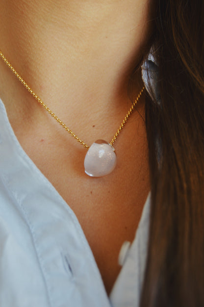 A close-up of a person wearing the Grand Bubble Single - Nude, a 14k gold filled chain necklace with a translucent stone pendant, against their skin and part of a light blue shirt.