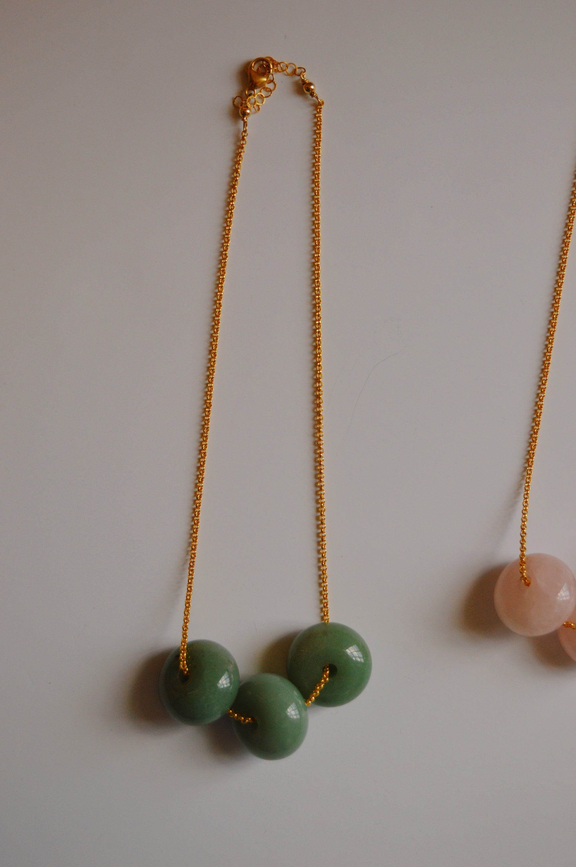 Grand Donut Layer-Green: A gold-filled chain necklace featuring three large green beads, with part of a pink beaded necklace visible on the right, displayed on a plain white surface—an elegant handcrafted jewelry piece.