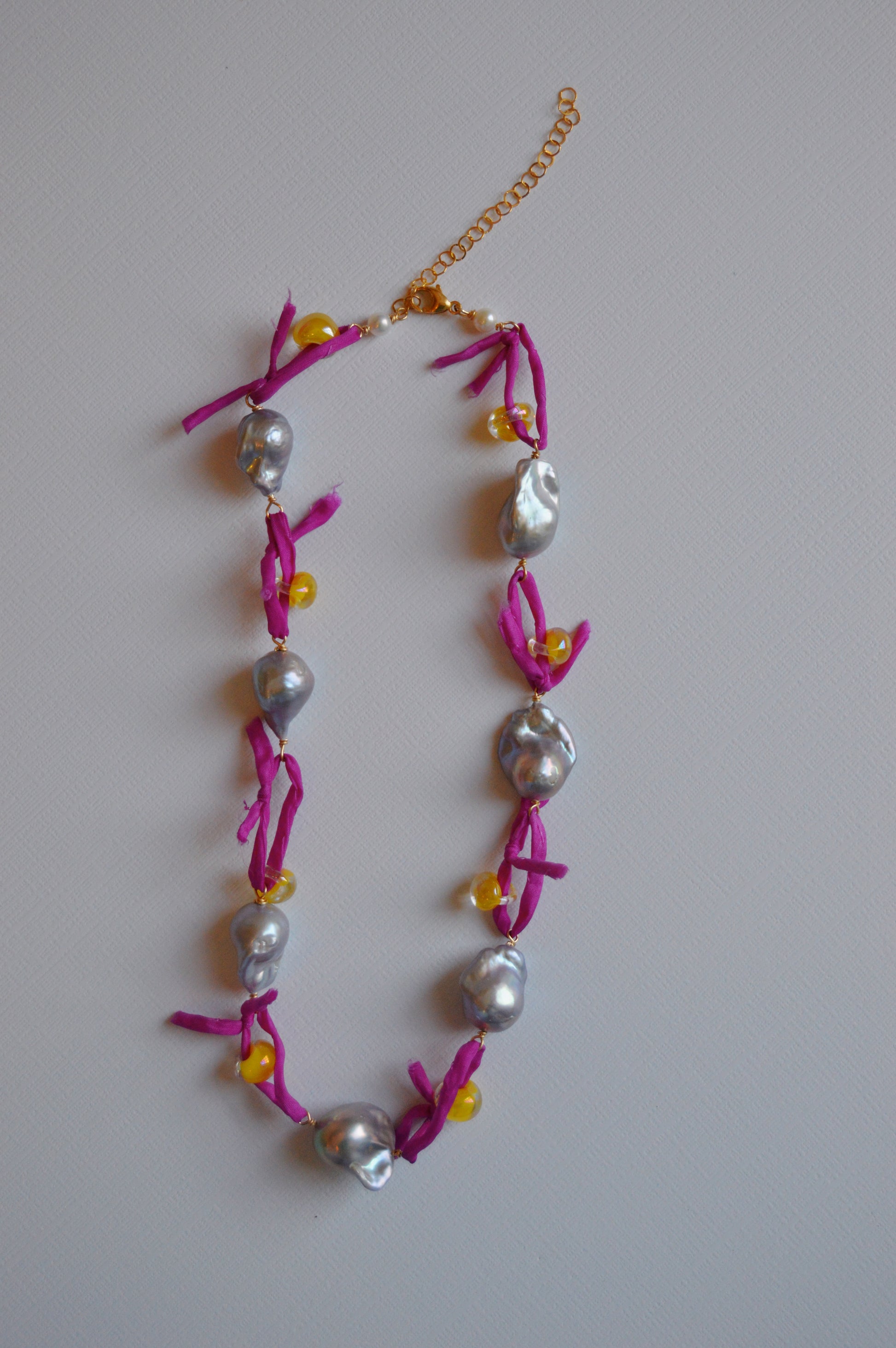 The Silk and Pearl Layer- Pink Lemonade Shimmer is a handmade necklace with irregular silver freshwater pearls, small yellow beads, and bright pink fabric knots on a gold adjustable chain, shown on a light gray surface.