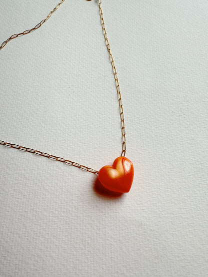 The Acrylic Heart Necklace features an orange acrylic heart pendant on a 14k gold filled chain, displayed on a textured white surface.