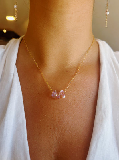 A close-up of a person wearing the Dainty Donut Layer, a 14K Gold Chain necklace featuring three small, translucent purple glass donut beads. The individual is dressed in white, with visible skin above the neckline.