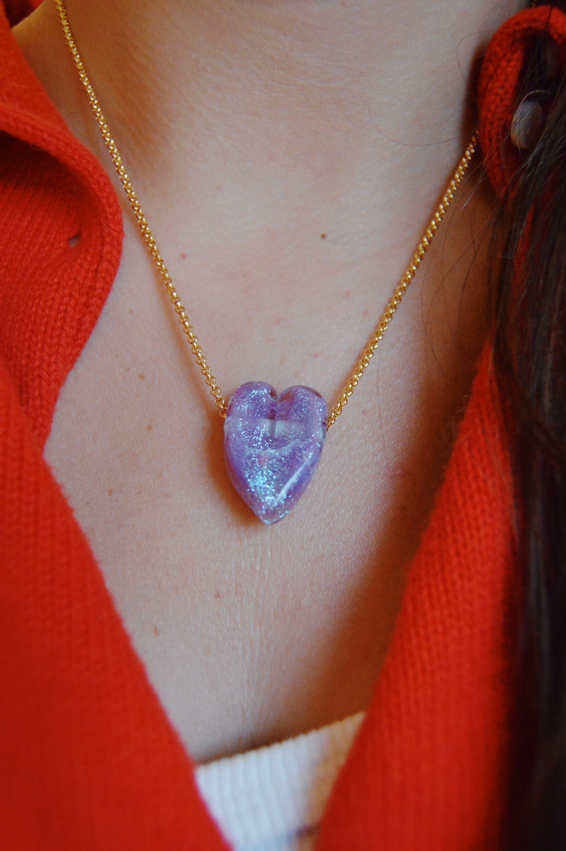 A close-up of someone wearing the Glass Heart Necklaces- Purples, a gold-filled chain with a handmade purple lampwork glass heart pendant, styled with a red shirt over a white top.