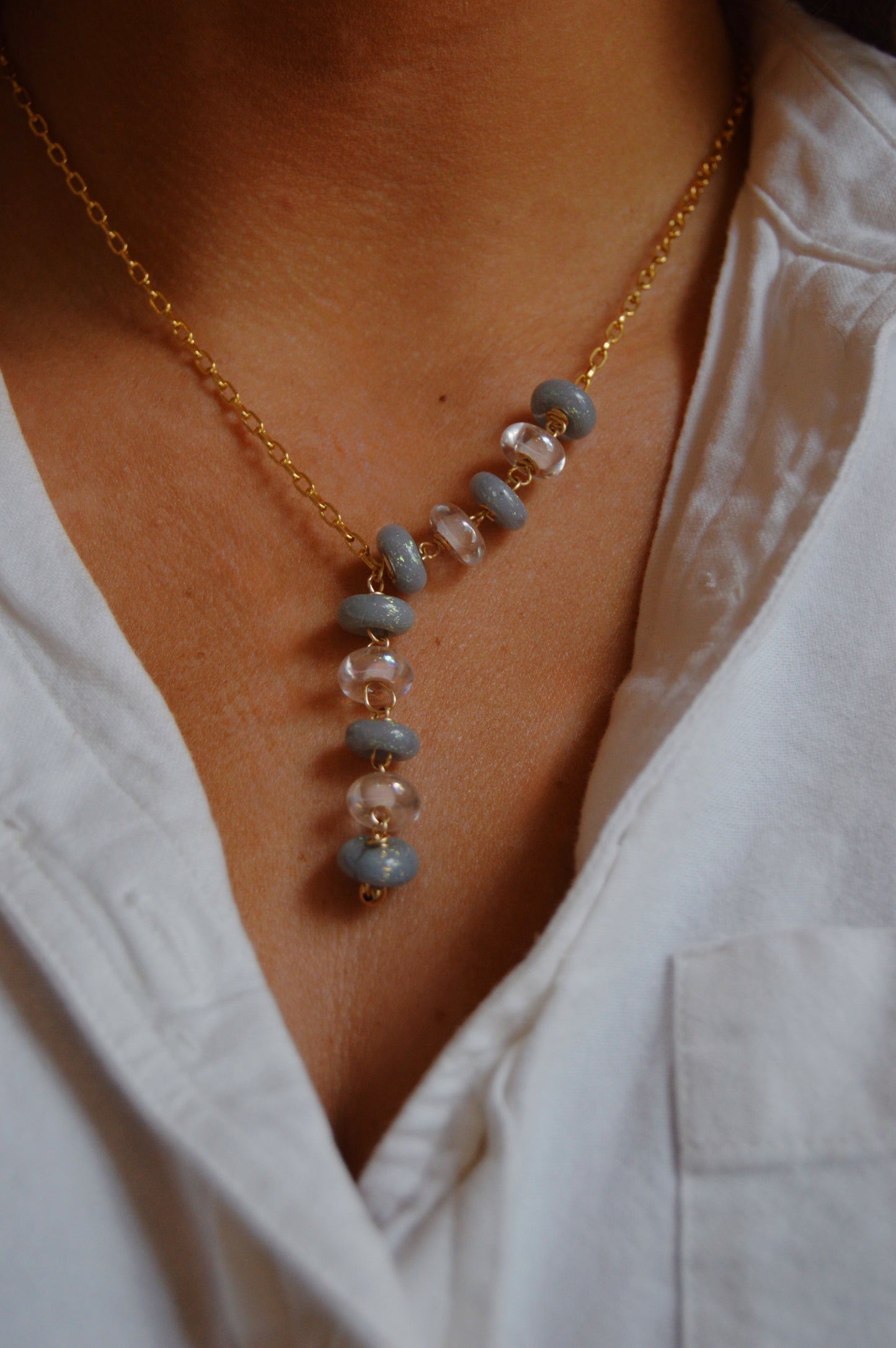 A person wears the Donut Lariat- Neutral Mix, a gold-filled chain necklace with alternating blue and clear lampwork glass beads, over a white collared shirt.