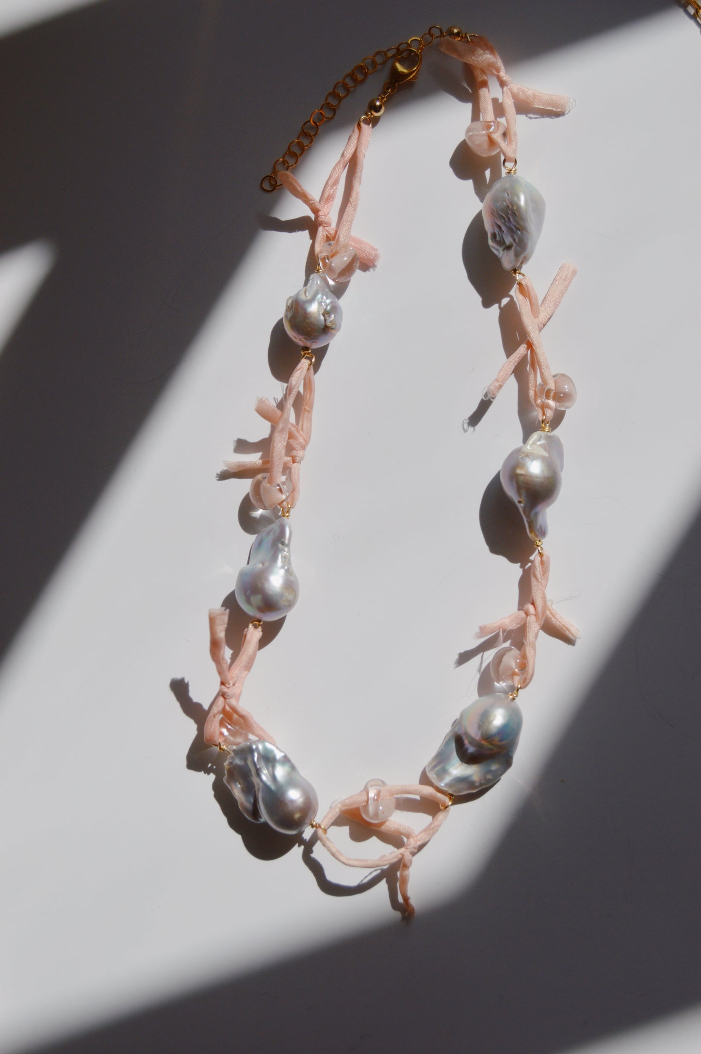 The Silk and Pearl Layer- Blossom necklace, showcasing large irregular freshwater pearls with pink ribbon accents, is displayed on a white surface with diagonal shadows highlighting this elegant handmade jewelry piece.
