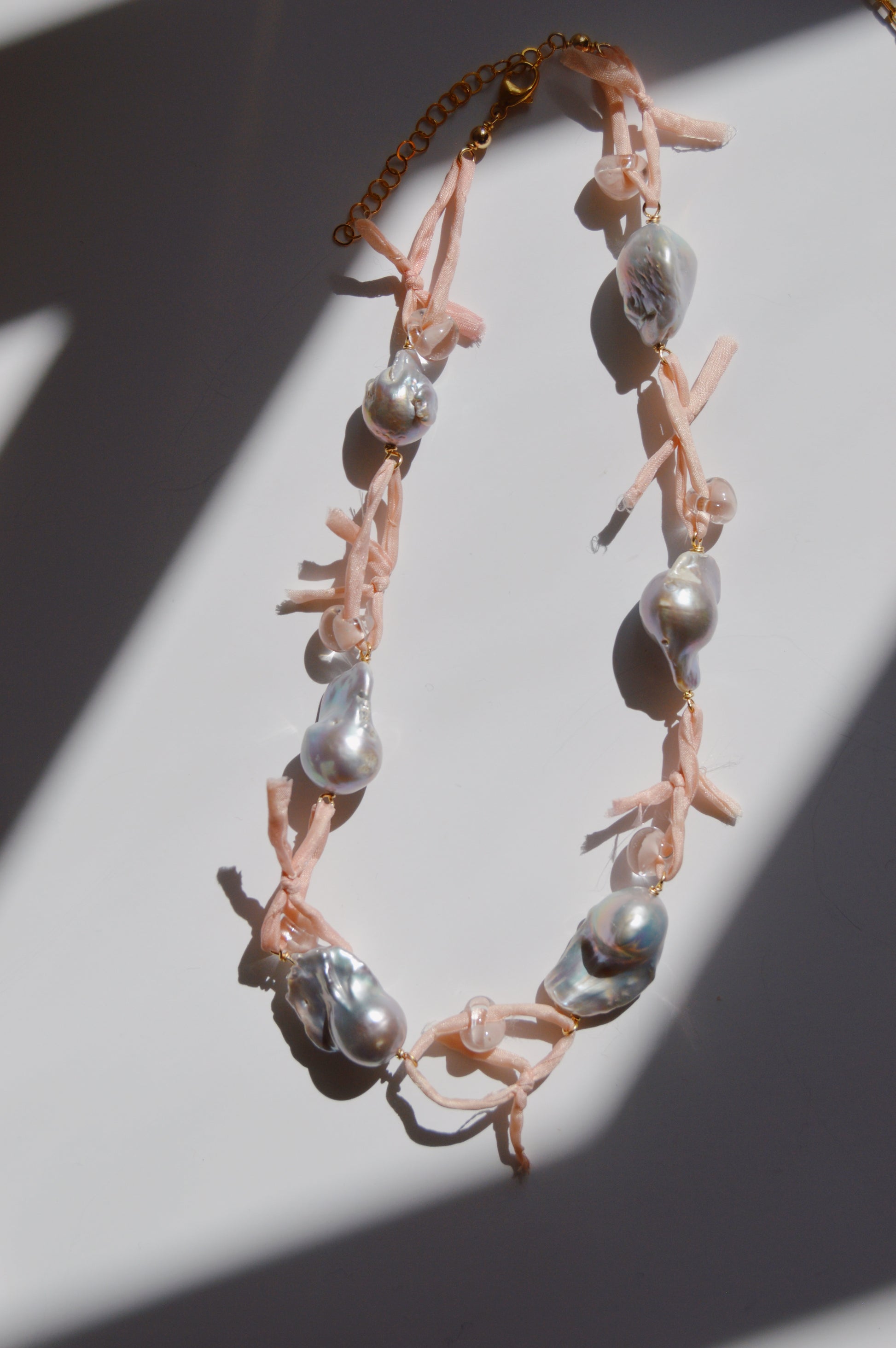 The Silk and Pearl Layer- Blossom necklace, showcasing large irregular freshwater pearls with pink ribbon accents, is displayed on a white surface with diagonal shadows highlighting this elegant handmade jewelry piece.