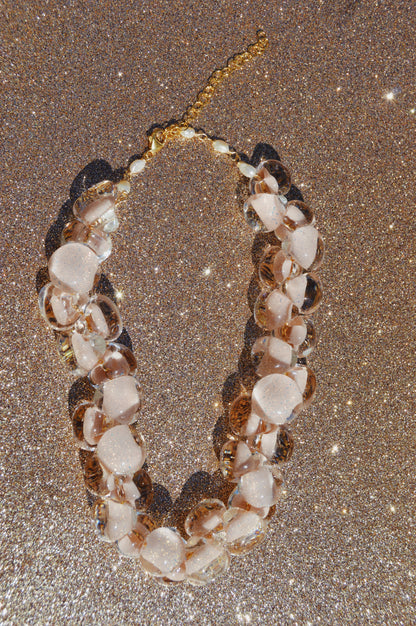 The Grand Bubble Glass Necklace-Nude features clear and pale pink glass beads, gold hardware, and an adjustable chain clasp, beautifully displayed on a glittery surface.
