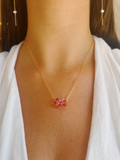 A woman in a white top wears the Dainty Donut Layer—a 14K gold chain necklace with three pink glass donut beads—paired with gold chain earrings.