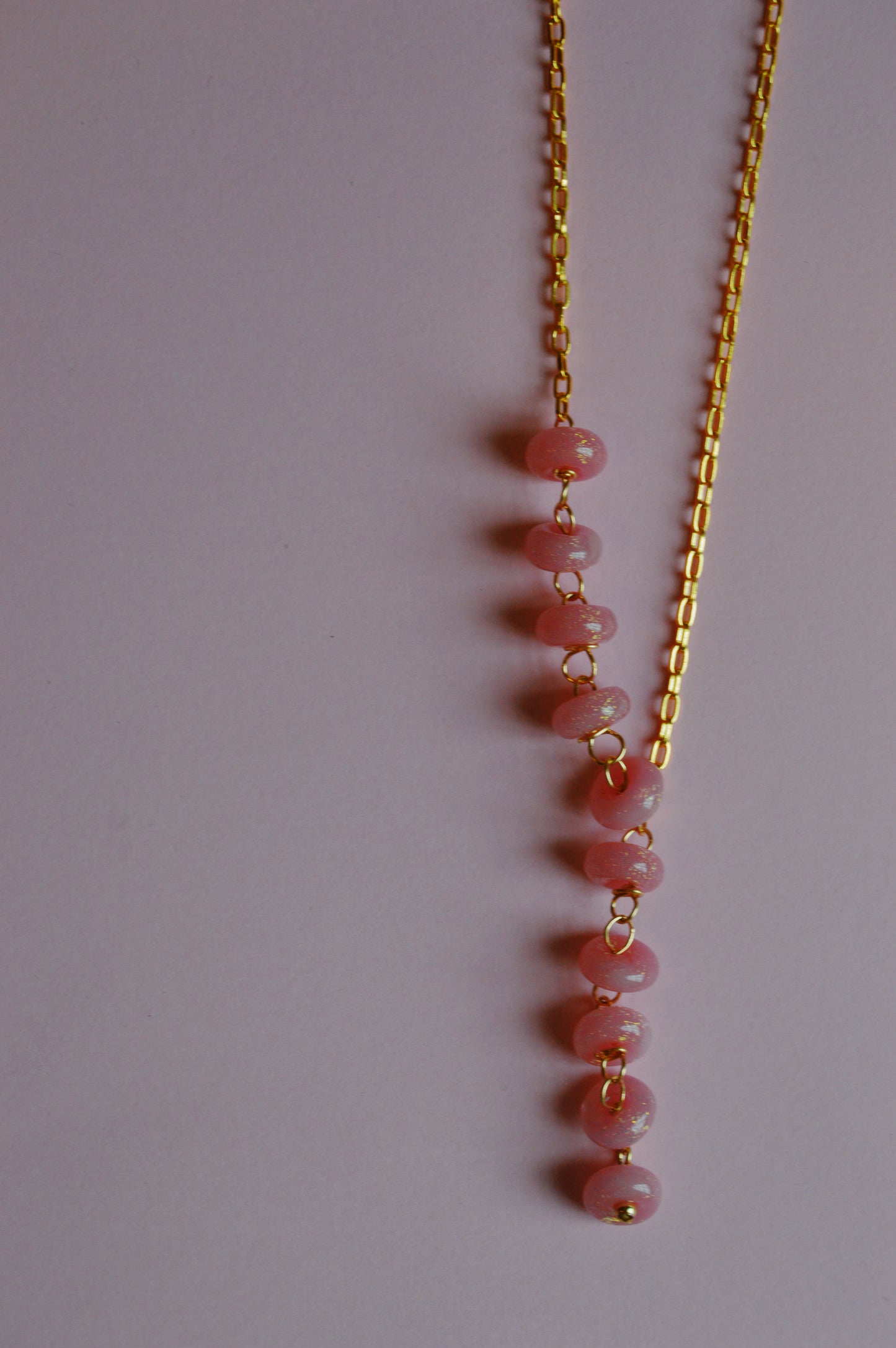 The Donut Lariat-Pink Sparkle is a gold chain necklace featuring a vertical row of pink beads, each separated by gold loops, displayed on a plain light pink background.