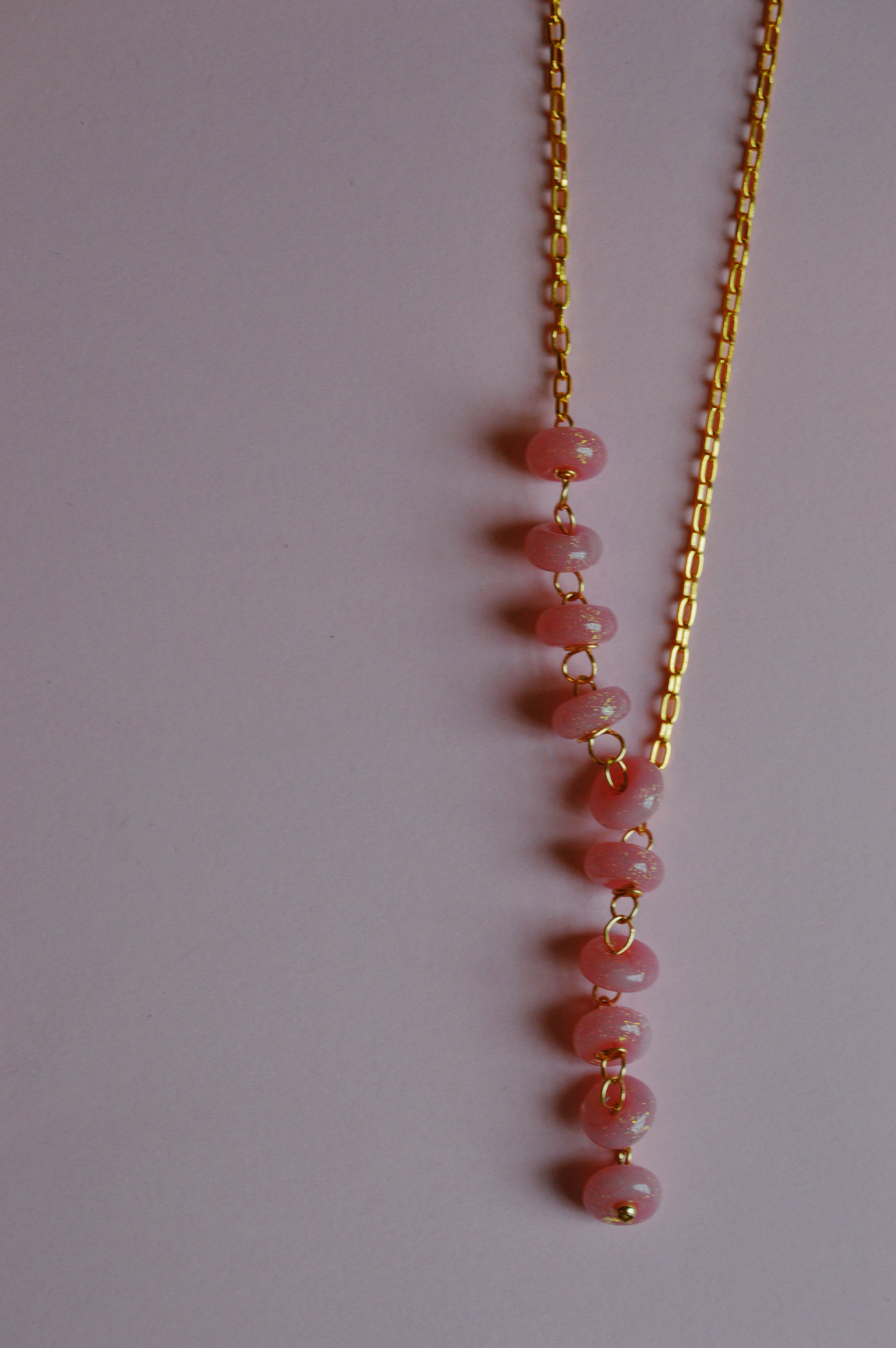 The Donut Lariat-Pink Sparkle is a gold chain necklace featuring a vertical row of pink beads, each separated by gold loops, displayed on a plain light pink background.