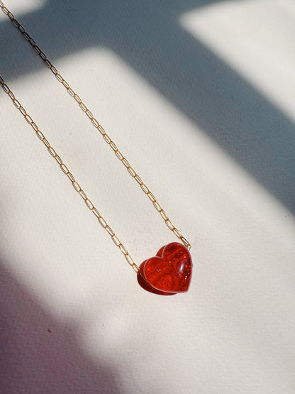 The Acrylic Heart Necklace, featuring a red acrylic heart pendant on a gold filled chain, rests on a white textured surface with soft patterns of light and shadow.