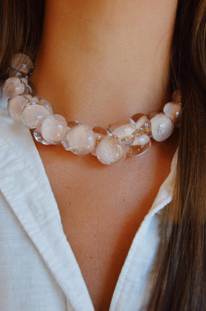 A close-up of a person wearing the Grand Bubble Glass Necklace-Nude—a chunky necklace of clear glass beads—over a white collared shirt, with long brown hair partially visible on both sides of their neck.