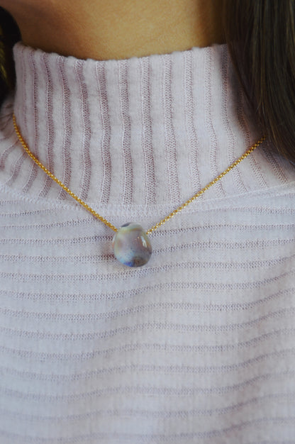 A close-up of a person wearing a ribbed, light purple turtleneck and the Grand Bubble Single - Jupiter: a gold filled chain necklace with a large, irregularly-shaped freshwater pearl pendant.