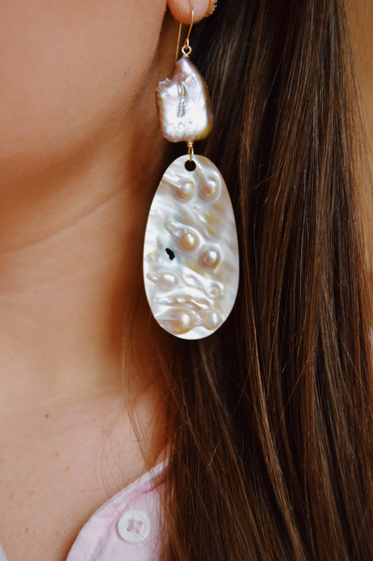 A close-up of a person wearing Angel Shell Drops, handcrafted earrings with large textured oval cultured freshwater pearls and a smaller irregular pearl above, all set on 14k gold-filled accents. Brown hair cascades beside the earring.