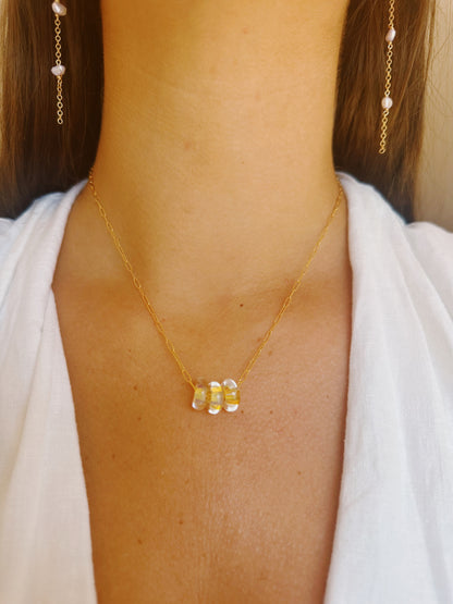 A close-up of a woman wearing the Dainty Donut Layer: a 14K gold chain necklace with three translucent stacked glass donut beads and matching gold chain earrings, paired with a white V-neck top.