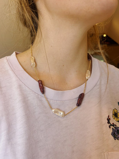 A person shown from the shoulders up wears the Saturday Layer, a 14K Gold Filled Chain necklace with alternating clear and dark red beads, paired with a light pink shirt featuring floral embroidery on the shoulder.