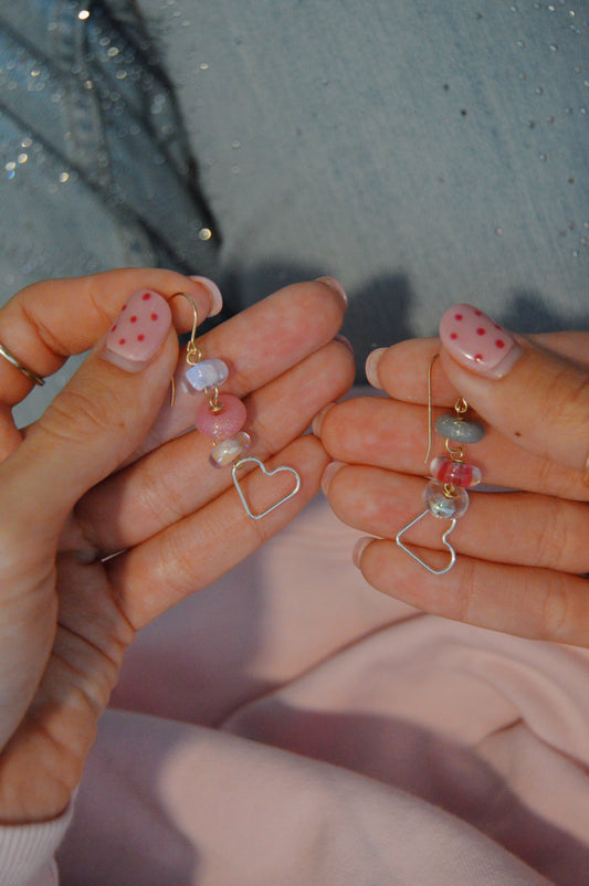 With pink and red polka dot nails, someone holds Silver Heart Drops—handcrafted dangling earrings with pastel beads and sterling silver heart charms—against a backdrop of a denim jacket and soft pink fabric.