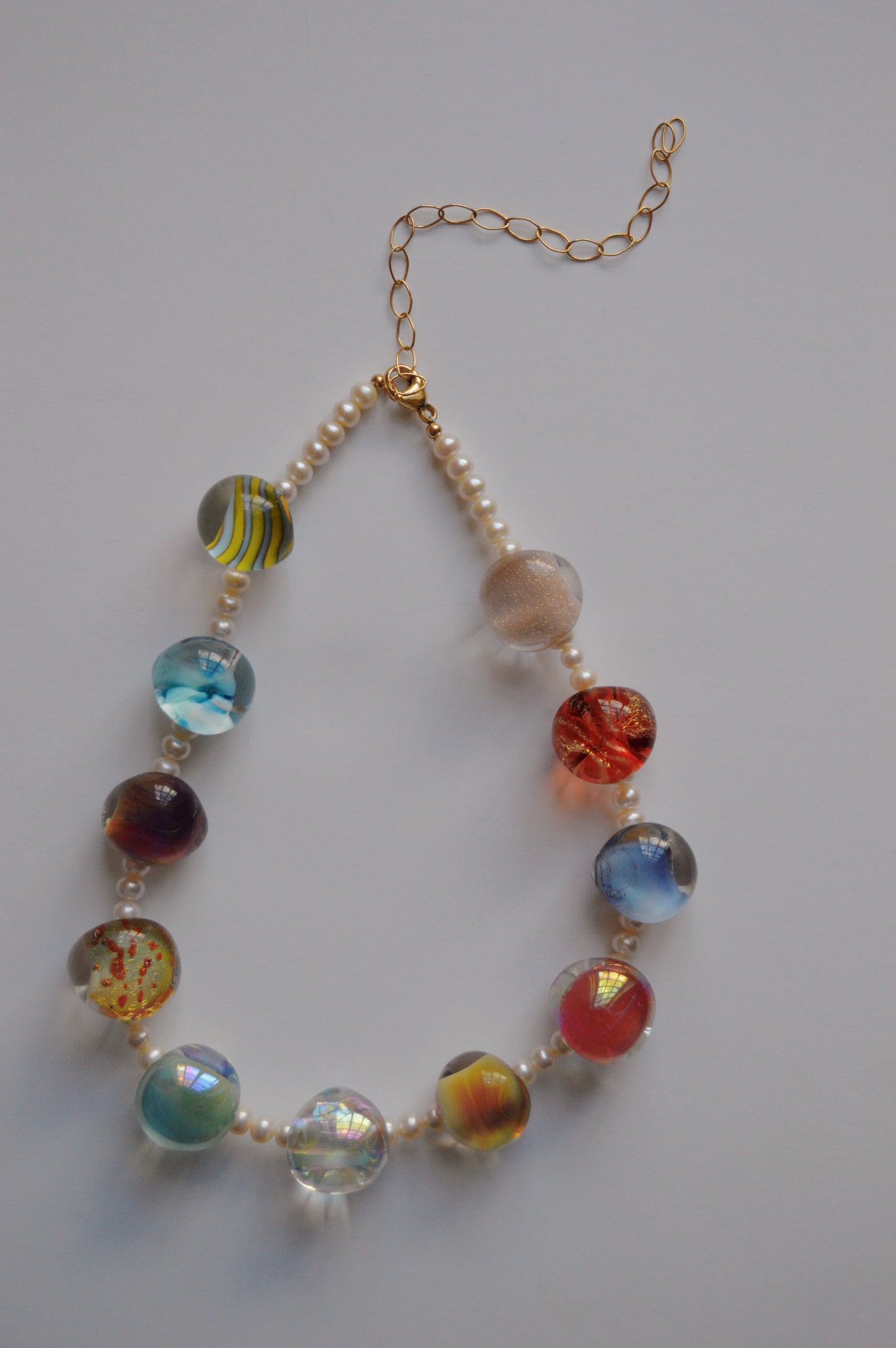 The Grand Bubble and Pearl Layer necklace features alternating handmade lampwork glass beads in various colors and patterns, small freshwater pearls, gold-filled findings, and a gold chain extender, shown on a white background.