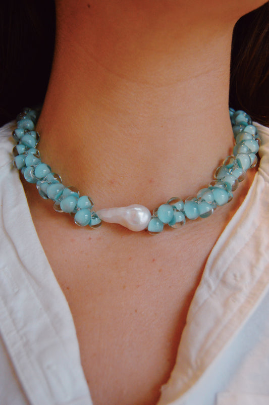 A person wearing a white shirt and the Bubble Glass Necklace- Santorini, a blue handmade glass bead necklace featuring a single large white pearl in the center.