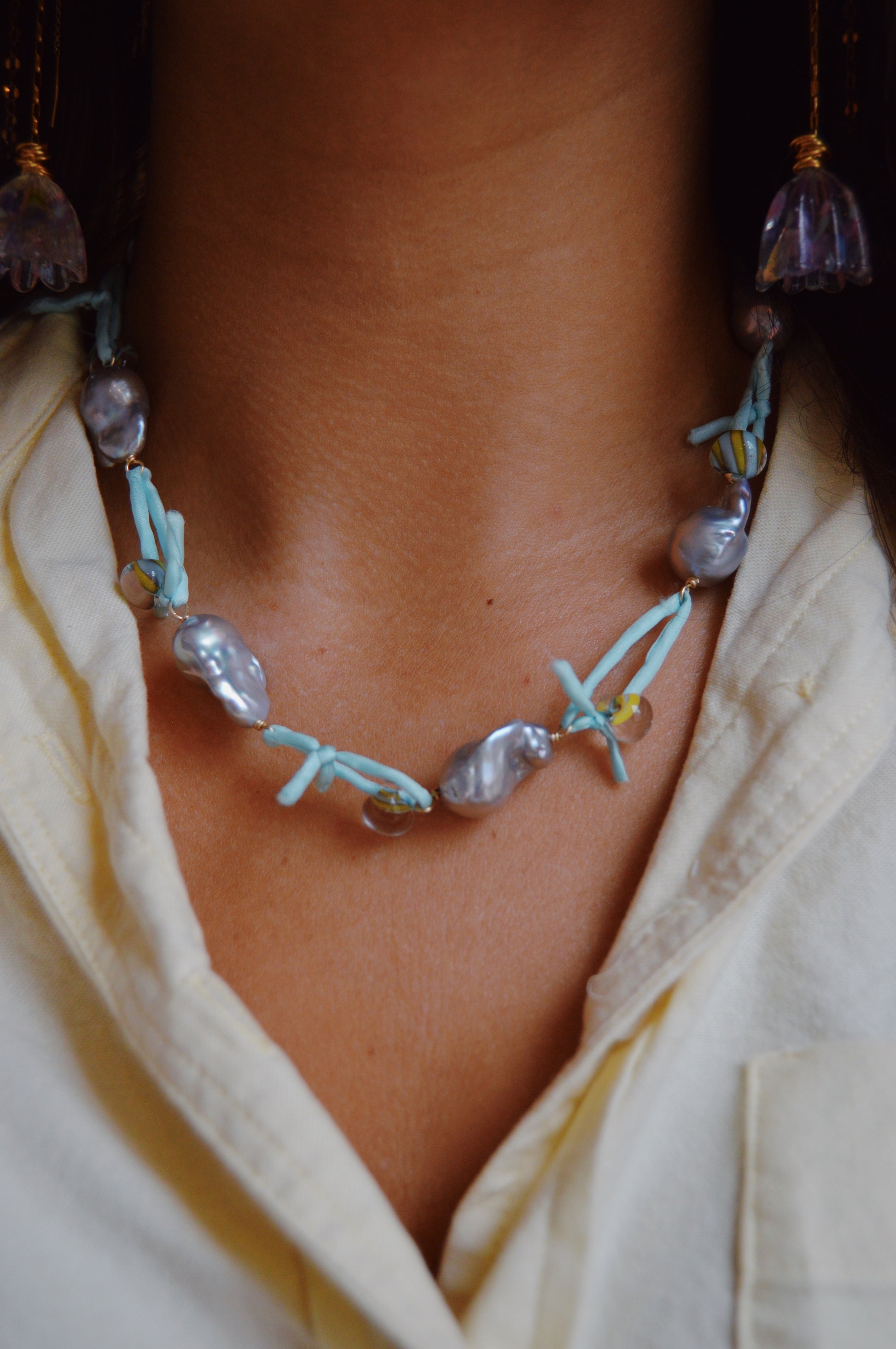 A close-up of a person wearing a pastel yellow shirt and the Silk and Pearl Layer- Robin's Egg, a handmade necklace featuring large irregular freshwater pearls, light blue beads, and small round beads resting on their neck and chest.