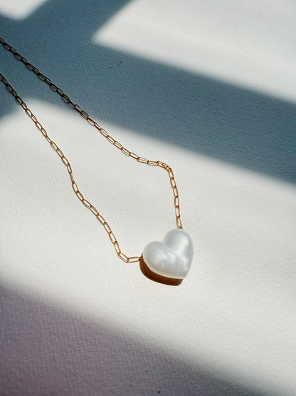 The Acrylic Heart Necklace, featuring a white acrylic heart pendant on a 14k gold filled chain, rests on a textured white surface bathed in soft natural light and intersecting shadows.