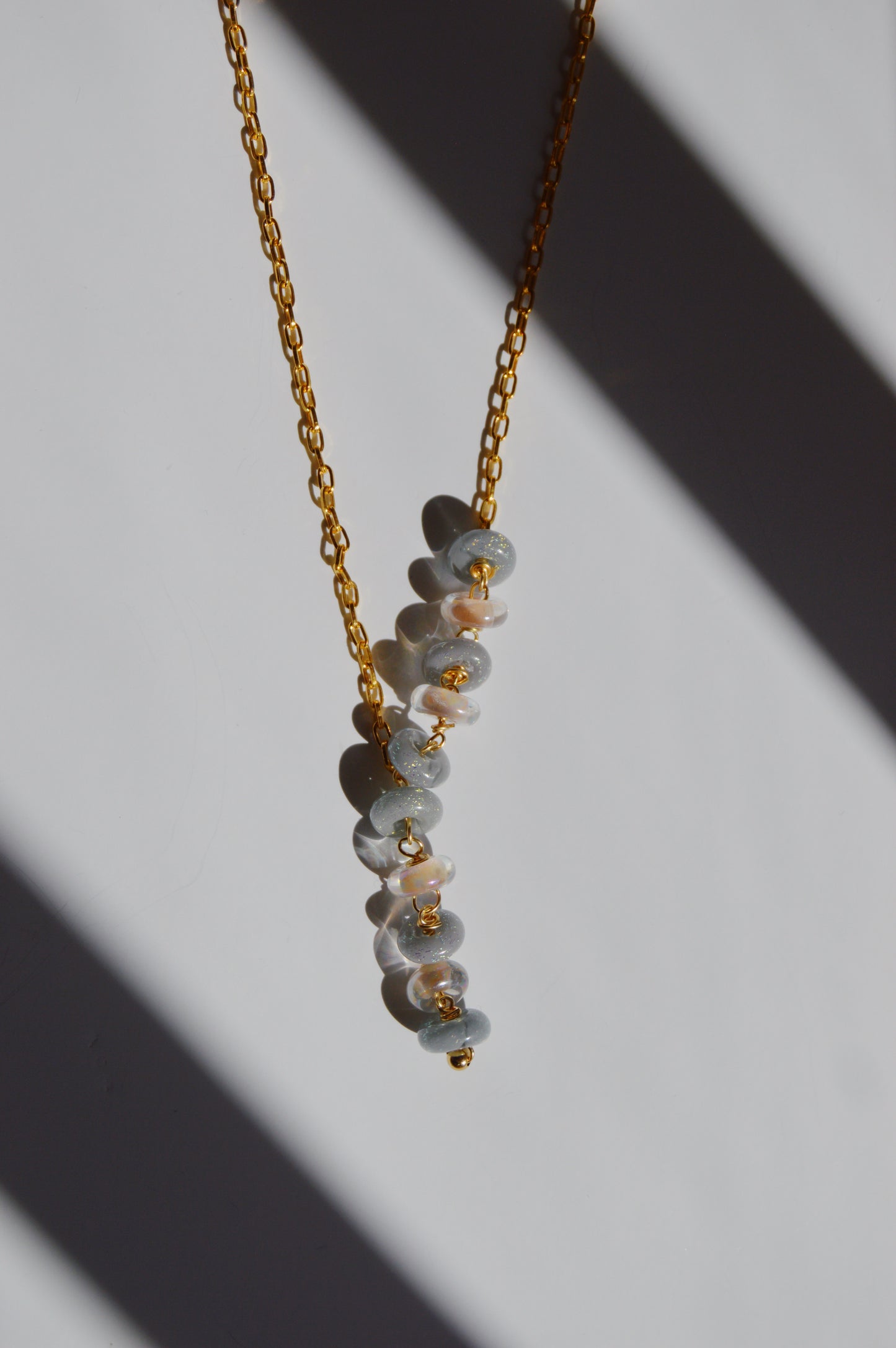 The Donut Lariat- Neutral Mix features stacked lampwork glass beads on a gold-filled chain, elegantly displayed on a white surface with diagonal shadows—an exquisite piece of handcrafted jewelry.