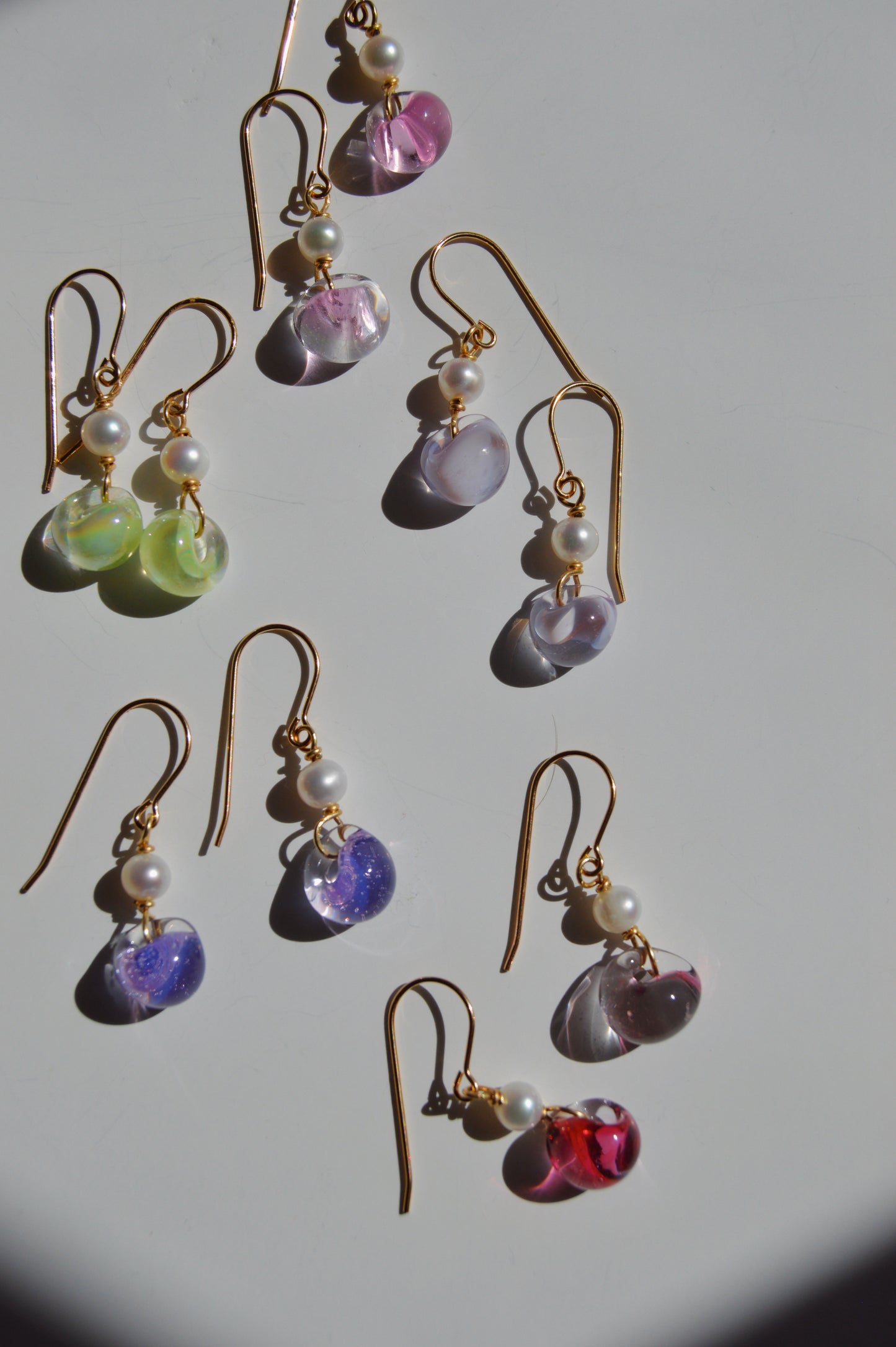 Nine pairs of Bubble Minis gold hook earrings with dangling glass beads in pink, green, purple, blue, and red rest on a white surface. Each features a small freshwater pearl and elegant gold filled findings.
