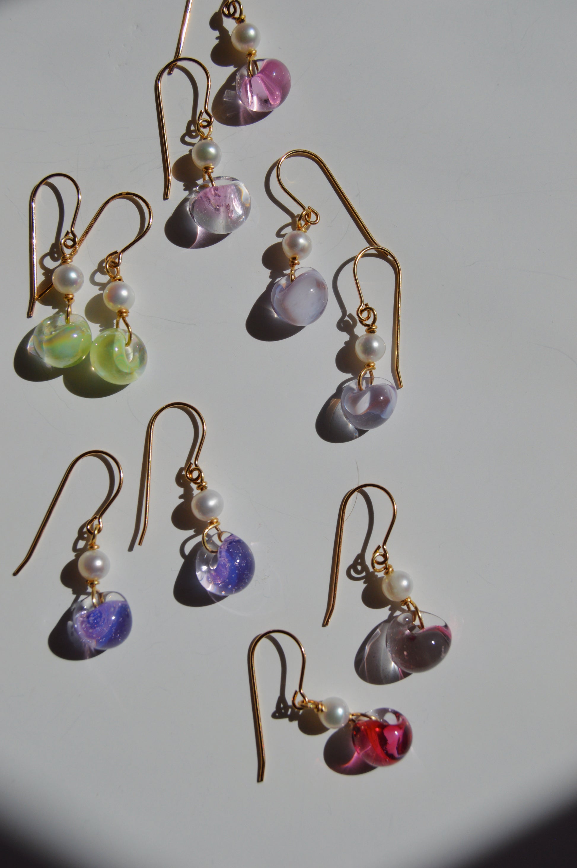 Nine pairs of Bubble Minis gold hook earrings with dangling glass beads in pink, green, purple, blue, and red rest on a white surface. Each features a small freshwater pearl and elegant gold filled findings.