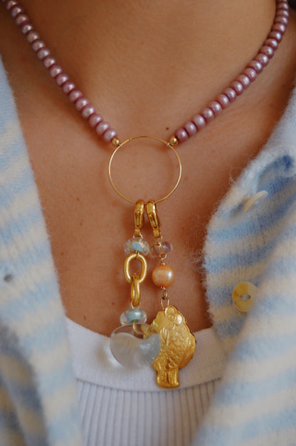 A person's neck wearing the Circle Pearl Layer necklace—a handcrafted purple beaded piece with a 14k gold-filled ring pendant, gold fish charm, Cultured Freshwater Pearls, and colored beads—paired with a white top and a blue-and-white striped cardigan.