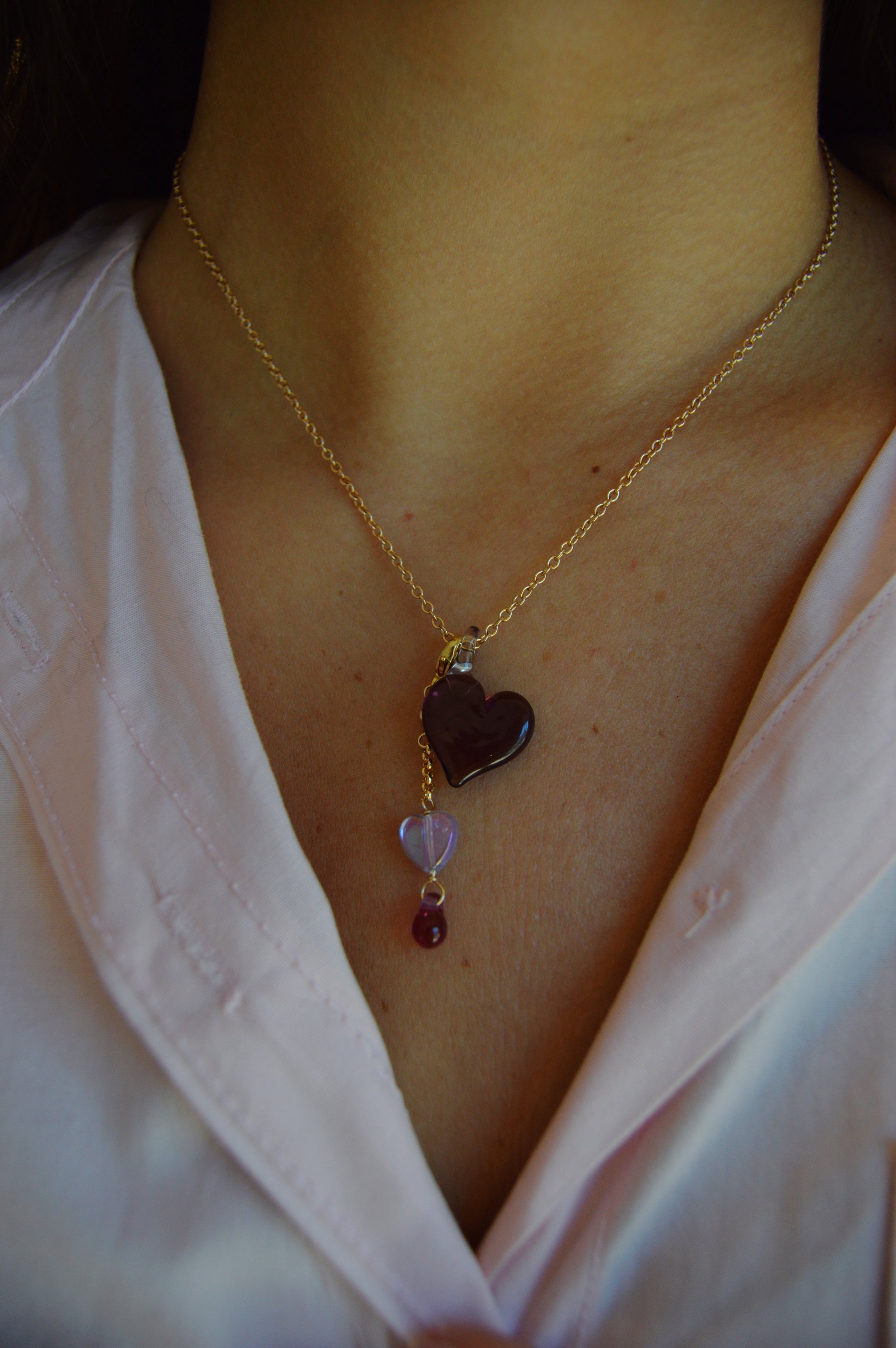 A person wears the Sweetheart Charm gold chain necklace, featuring a dark heart, a small purple heart, and a round red bead pendant, over a pale pink collared shirt.