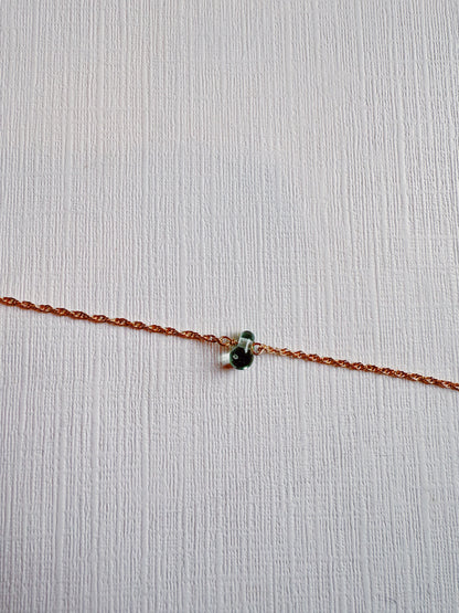 The Dewdrop Bracelet features a delicate 14k gold filled chain with a small dark green bead charm, shown diagonally on a white textured surface.