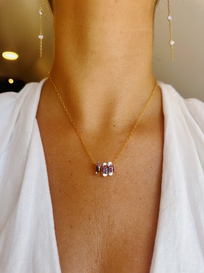 A close-up of a person wearing the Dainty Donut Layer— a 14K gold chain necklace featuring three purple and silver glass donut beads—paired with dangling gold earrings and a white v-neck top.