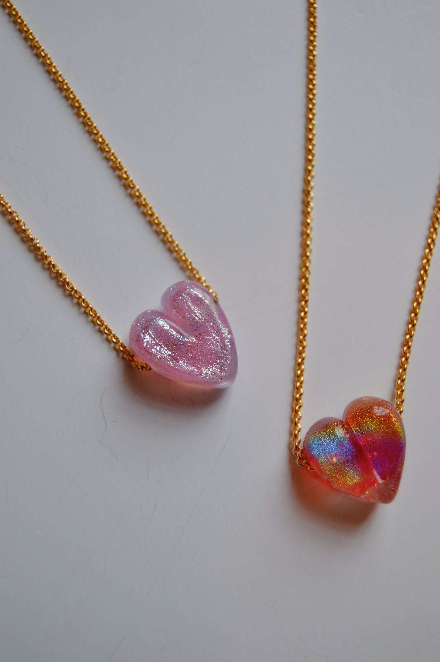 Two Glass Heart Necklaces- Pinks—featuring gold-filled chains and handmade lampwork glass heart pendants in shimmery pink and iridescent red-blue hues—rest on a white surface, highlighting artisan jewelry craftsmanship.